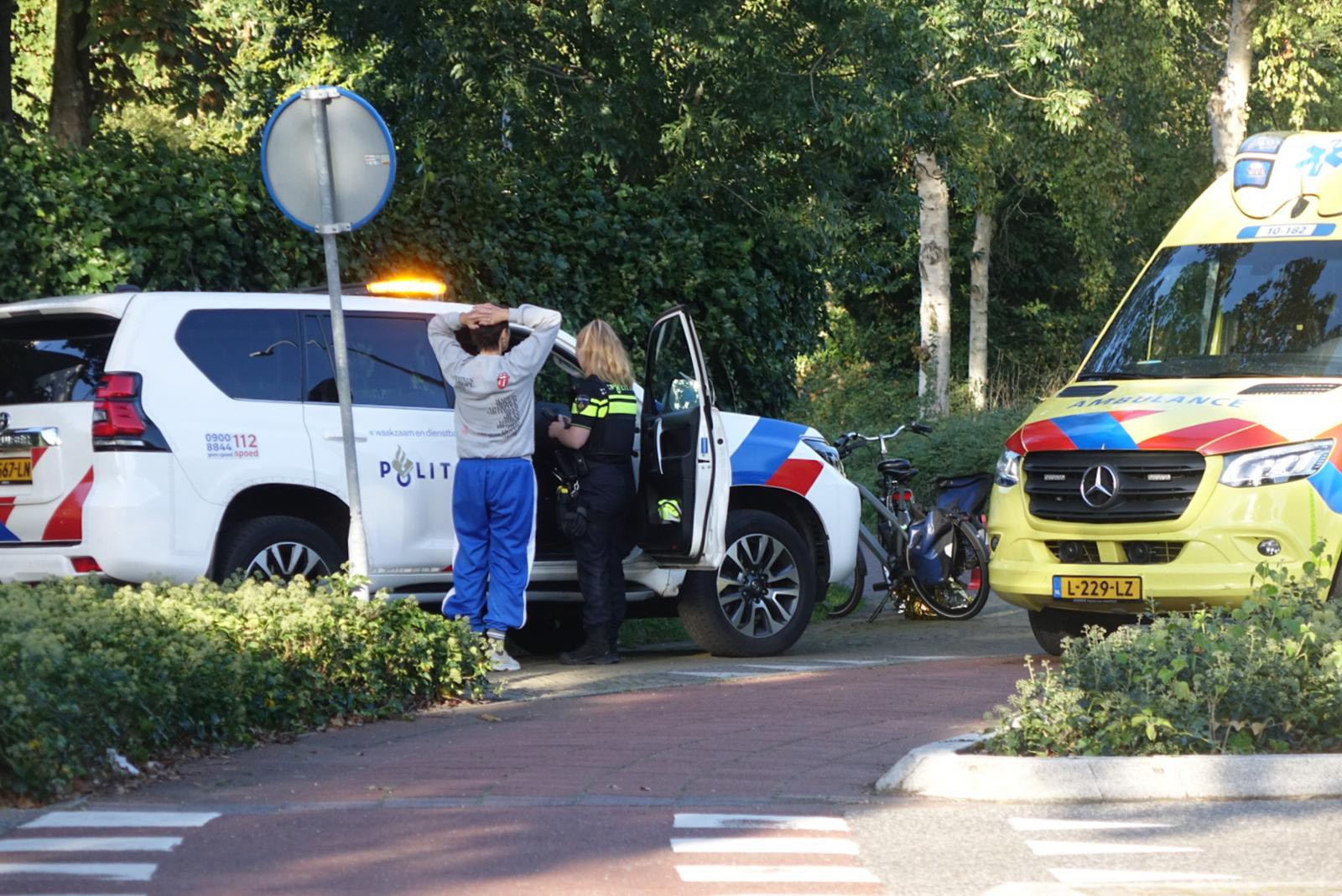Ambulance geparkeerd langs de weg terwijl hulpverleners een slachtoffer behandelen; omstanders met fietsen kijken toe