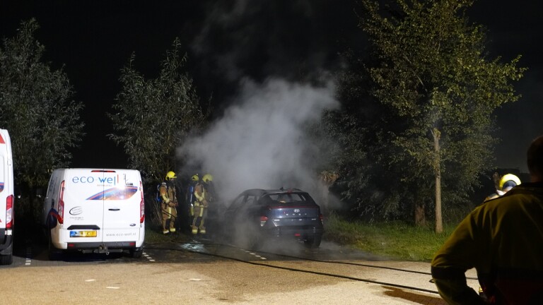 Brandweerman blust een brandende auto met grote vlammen en rook op een donkere weg naast een bestelbus
