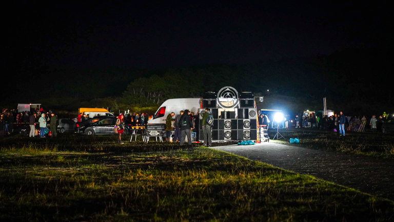 Nachtelijke buitenbijeenkomst met mensen rond een grote speakerset op een aanhanger, met busjes en felle lampen