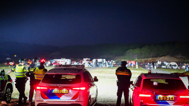 Nachtelijke buitenbijeenkomst met mensen rond een grote speakerset op een aanhanger, met busjes en felle lampen