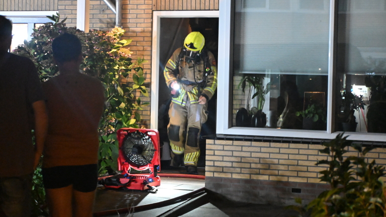 Brandweerman in volledige uitrusting loopt met zaklamp uit een woning, met ventilator en brandslangen bij de deur