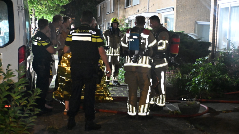 Brandweerman in volledige uitrusting loopt met zaklamp uit een woning, met ventilator en brandslangen bij de deur
