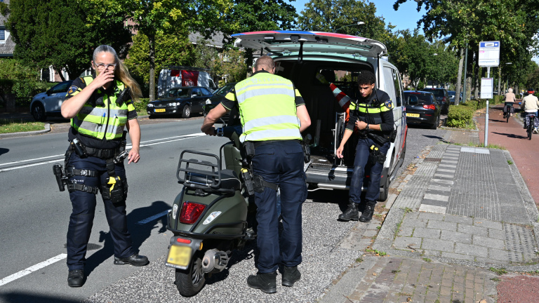 Ambulance en politie bij een stilstaande auto; hulpverleners praten met een vrouw naast een scooter in woonwijk