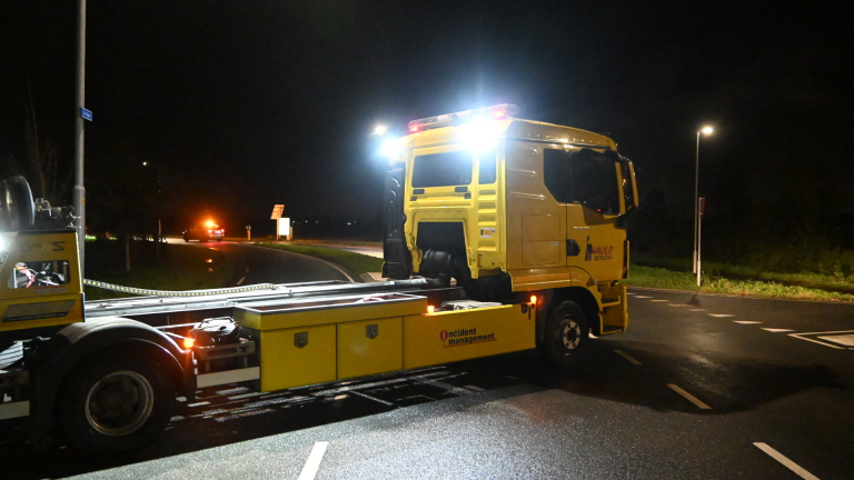 Auto in de berm langs een sloot, met een hulpverlener in oranje reflectiekleding bij het voertuig in de nacht