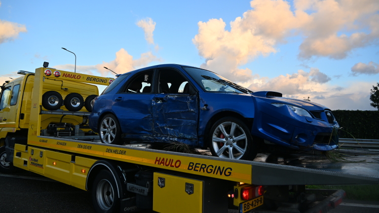 Blauwe auto met schade aan de zijkant staat deels in de berm naast een weg, omringd door gras en struiken