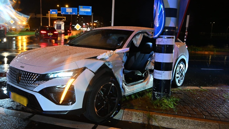 Zwaar beschadigde auto op nat wegdek na aanrijding, met ambulance en hulpverleners op de achtergrond