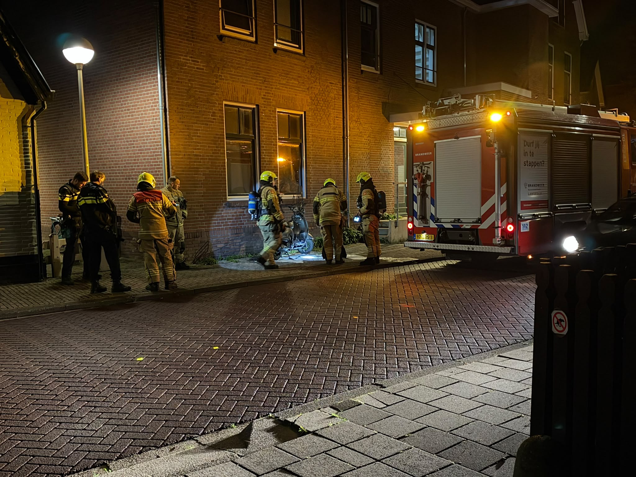 Brandweerlieden bij een bakstenen gebouw naast een brandweerwagen, inspecteren een uitgebrande scooter op straat