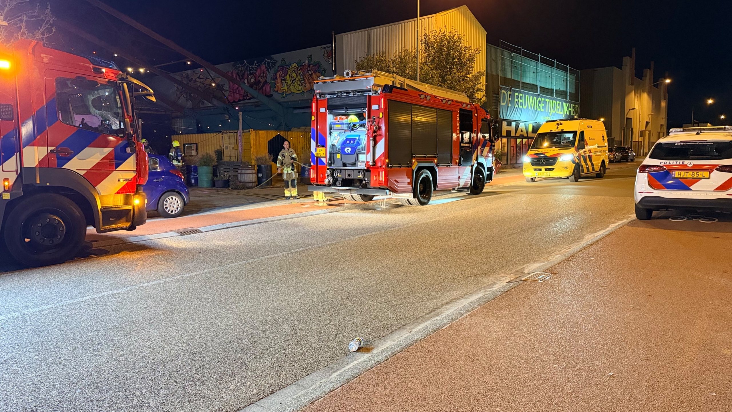 Brandweerlieden staan ’s avonds bij een deels uitgebrande loods met gebroken ruiten en een groot graffiti-paneel ervoor