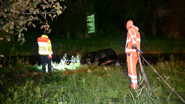 Zwarte auto deels in een sloot beland langs een grasberm, met modder op de carrosserie en koplampen aan