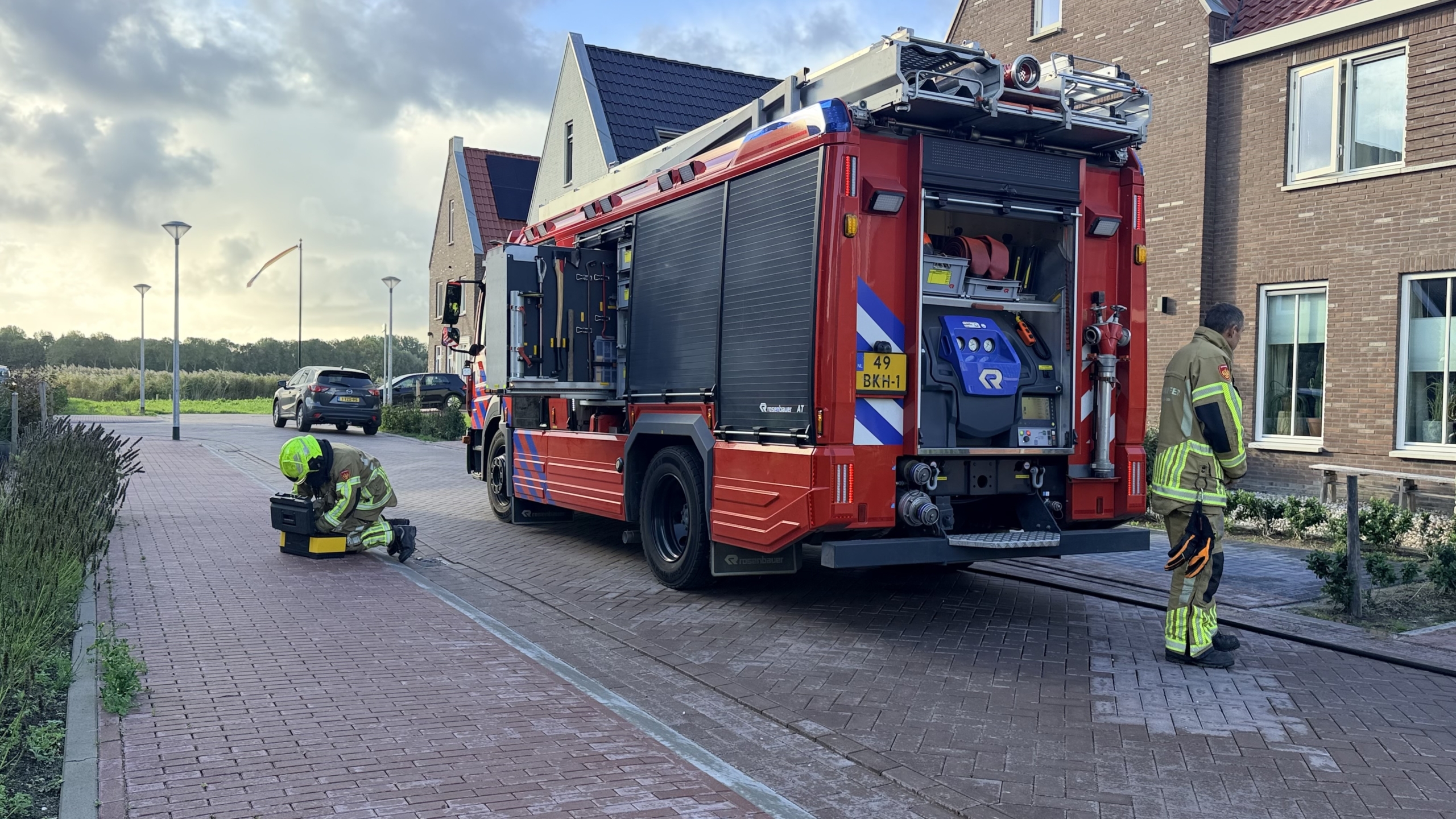 Brandweer bij auto met open motorkap op oprit; brandweerwagen en slangen in woonstraat, omstanders erbij