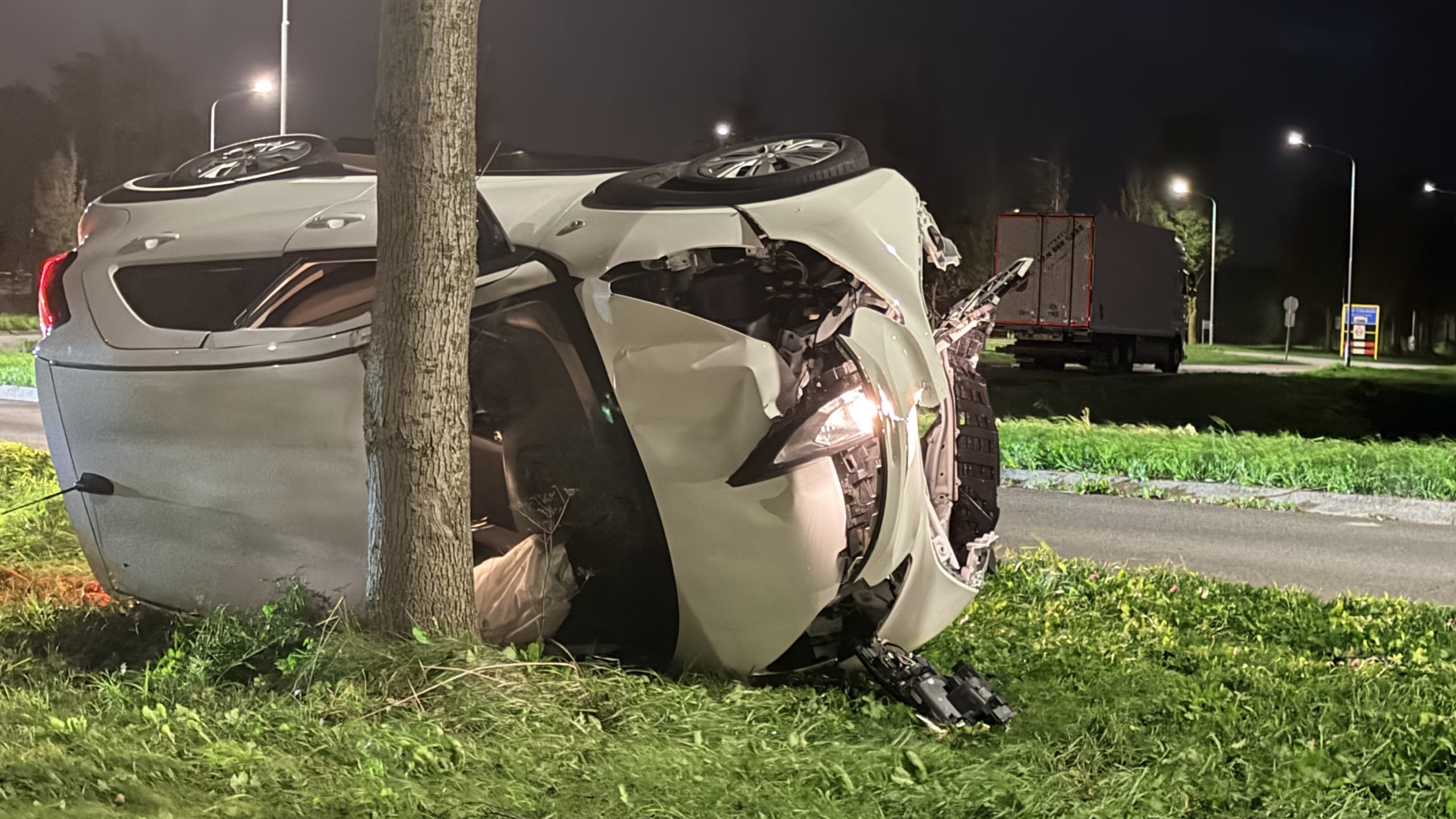 Omgeslagen auto tegen een boom op een grasveld bij nacht, met politieagent en hulpverlener in de buurt