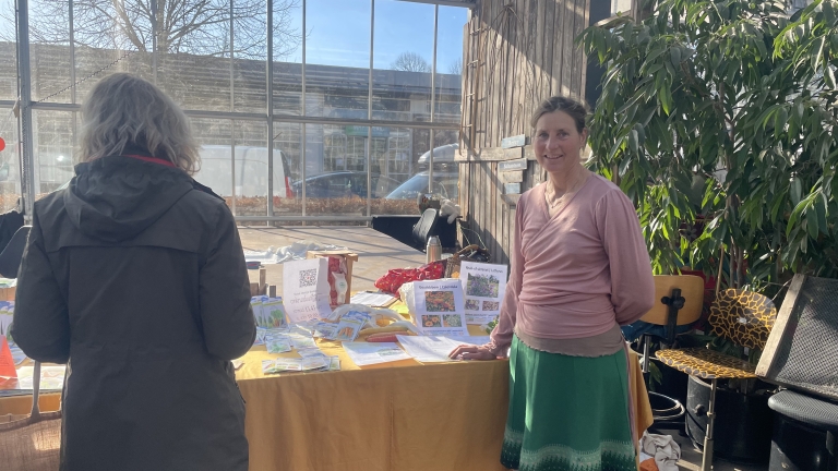 Vrouw staat bij een tafel met brochures en potten in een zonnige ruimte met grote planten en een glazen wand.
