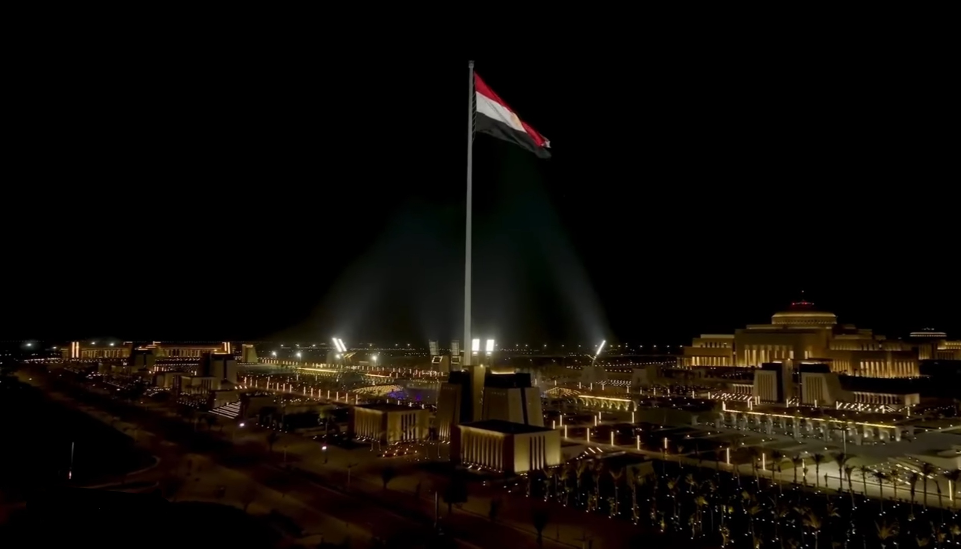 Nachtfoto van een groot, verlicht gebouw met een hoge vlaggenmast en een wapperende vlag, omringd door een uitgestrekt, verlicht terrein.