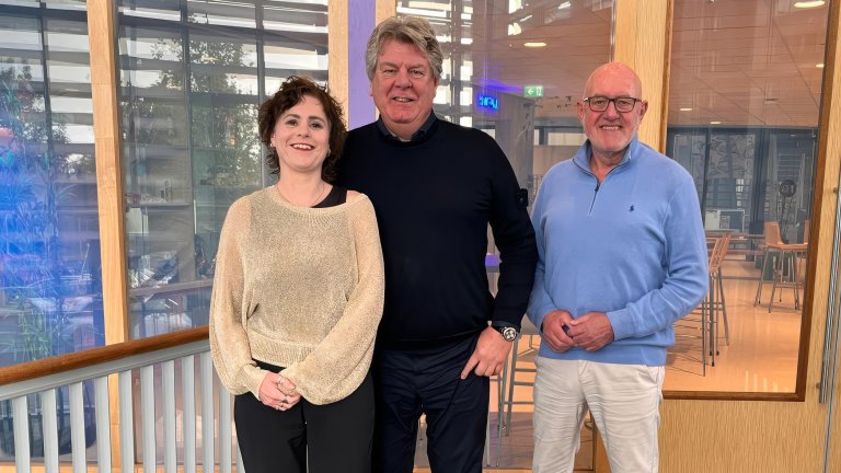 Drie glimlachende collega’s poseren in een modern kantoor met glazen wanden en hoge tafels op de achtergrond