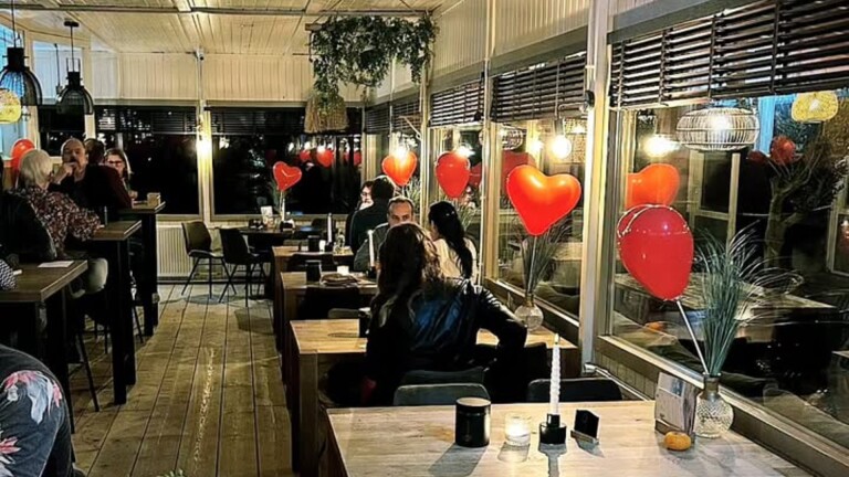 Een gezellig restaurant met houten vloeren en tafels versierd met rode hartvormige ballonnen en kaarsen. Er zitten enkele mensen aan hun tafels, pratend en genietend van de sfeer. Er hangen planten en lampen aan het plafond en het interieur is warm en uitnodigend.