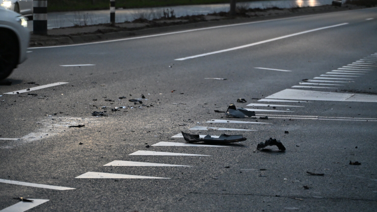 Ravage van autowrakken verspreid over een verlaten weg na een ongeval, met een langsrijdende auto op de achtergrond.