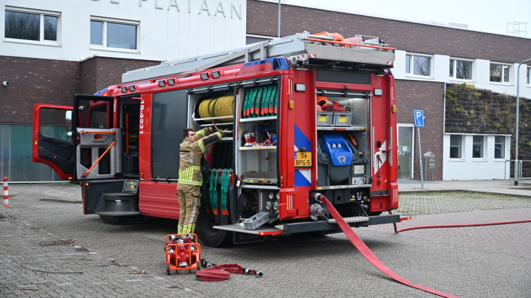 Brandweerman haalt materiaal uit geopende brandweerwagen voor een gebouw met het bord "DE PLATAAN".