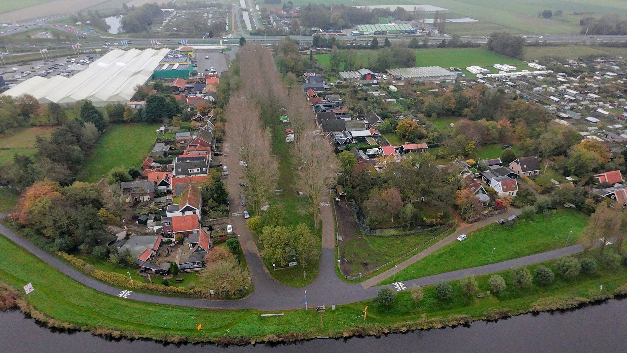 Luchtfoto van een klein dorp met huizen en een laan met bomen, omgeven door weilanden en een waterweg op de voorgrond. Op de achtergrond zijn kassen en een parkeerplaats zichtbaar.