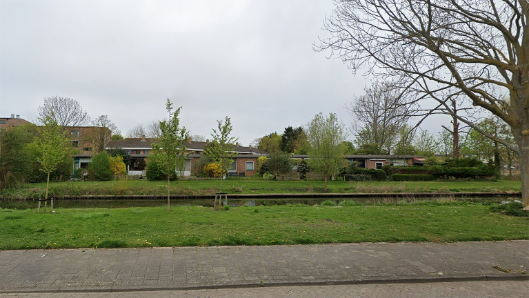 Lage huizen en bomen langs een smal waterkanaal, met een grasveld en een trottoir in de voorgrond.
