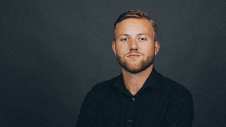 Man met kort, lichtbruin haar en een baard, gekleed in een donker overhemd, poserend tegen een donkere achtergrond.
