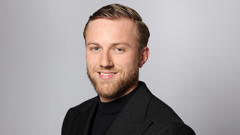 Man met baard en kort, gestyled haar in een zwart pak, glimlachend voor een grijze achtergrond.