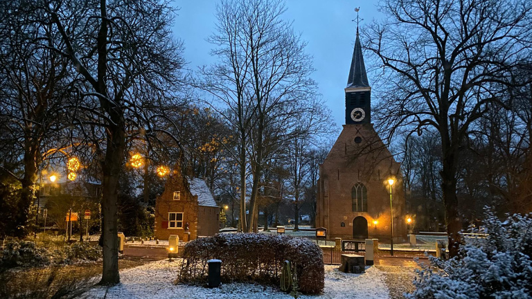 Besneeuwde kerk en omgeving bij schemering, met verlichting en kale bomen.