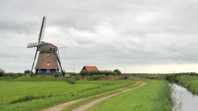 Molen in een groen landschap met een onverharde weg en een sloot onder een bewolkte hemel.