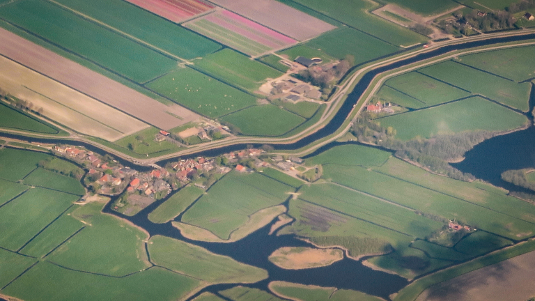 Luchtfoto van een Nederlands landschap met groene velden, enkele huizen en een kanaal dat door het gebied loopt, omringd door wegen en landerijen.