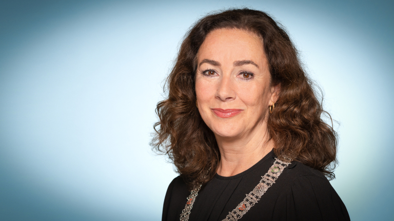 Portret van een vrouw met halflang krullend bruin haar, in zwarte top met decoratieve schouderband, tegen lichtblauwe achtergrond
