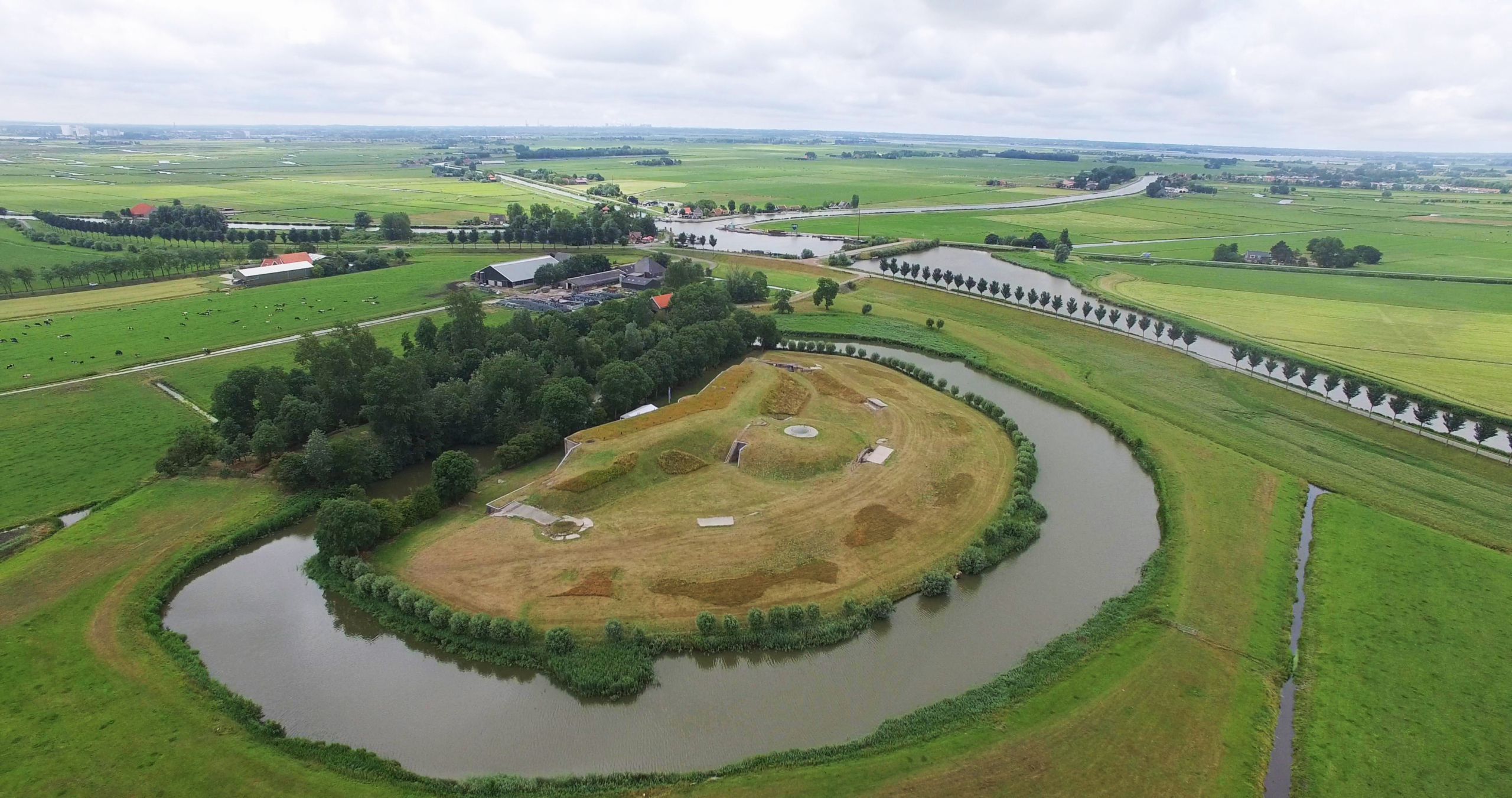 Luchtfoto van een groen polderlandschap met een rond fort omringd door water en omgeven door bomen en weide.