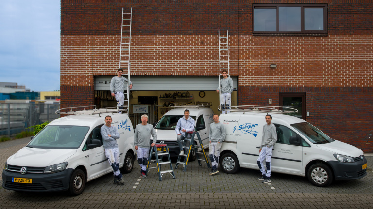 Vijf mannen poseren voor en op twee witte bestelwagens met ladders, tegen de achtergrond van een bakstenen gebouw.