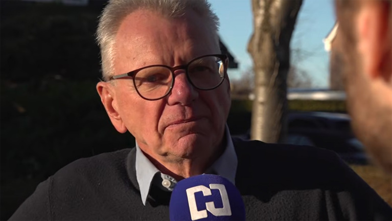 Een oudere man met een bril wordt buiten geïnterviewd en houdt een microfoon vast met een C-logo.