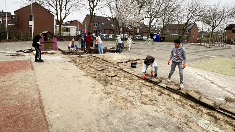 Kinderen op een schoolplein zijn bezig met het loshalen van stenen en hout. Ze dragen handschoenen en werken samen aan een project naast een speelhuisje. Op de achtergrond staan bomen en woonhuizen.