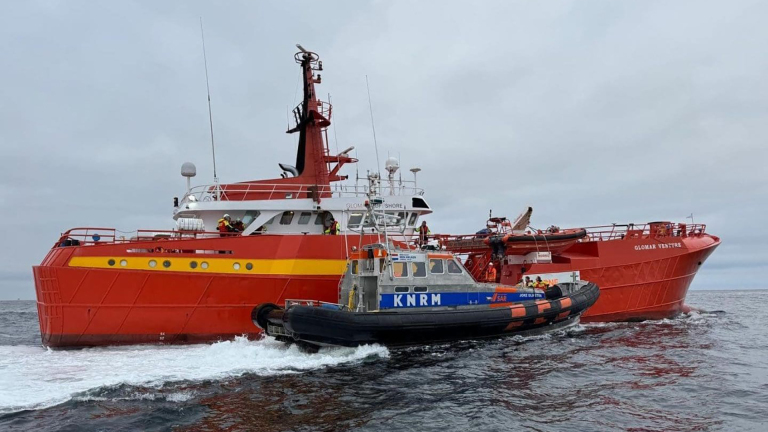 Een grote rode reddingsboot, "Glomar Venture," vaart naast een kleinere KNRM-boot op zee.