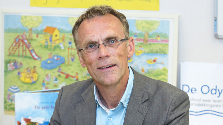 Man met bril en grijs haar draagt een grijs pak en lichtblauw overhemd, zittend voor een kleurrijk schoolbord met afbeelding van een speeltuin.