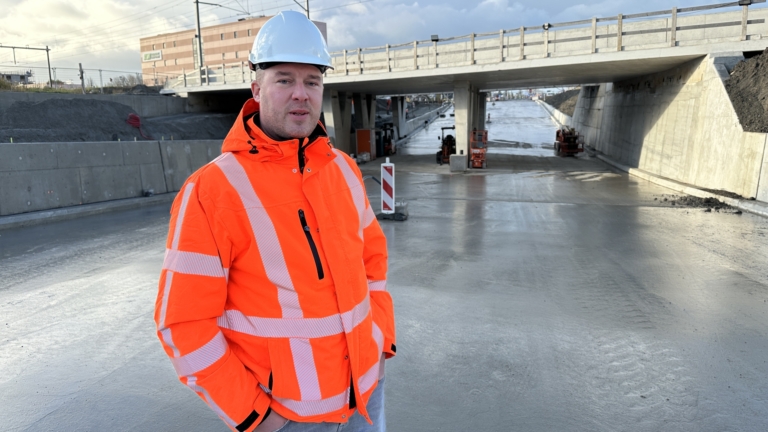 Een man in een oranje veiligheidshesje en witte helm staat op een bouwplaats onder een brug, met apparatuur op de achtergrond en een blauwe lucht vol wolken.