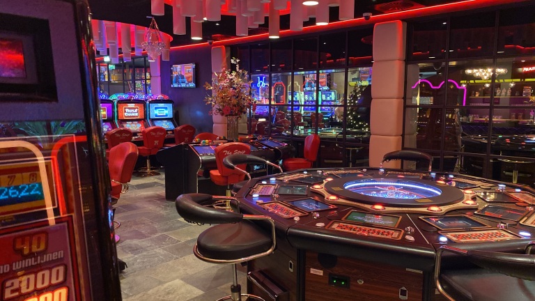 Een casino-interieur met speelautomaten, rode stoelen en een grote speeltafel met schermen en verlichting.