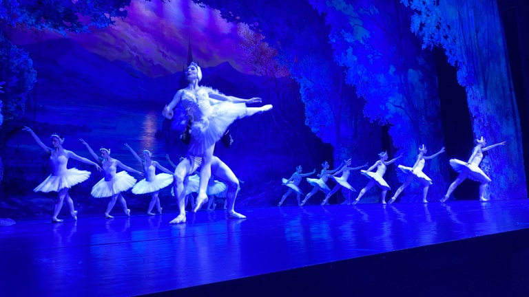 Balletdansers in witte tutu's voeren een pirouette uit op een podium met een blauw verlicht bos als achtergrond.