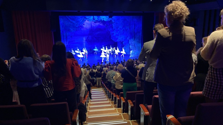 Publiek geeft staande ovatie in een theater, terwijl balletdansers op het podium buigen voor een blauw verlicht decor.