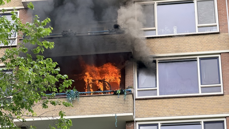 Appartementbrand met vlammen en rook op een balkon van een bakstenen gebouw, met groene bladeren op de voorgrond.