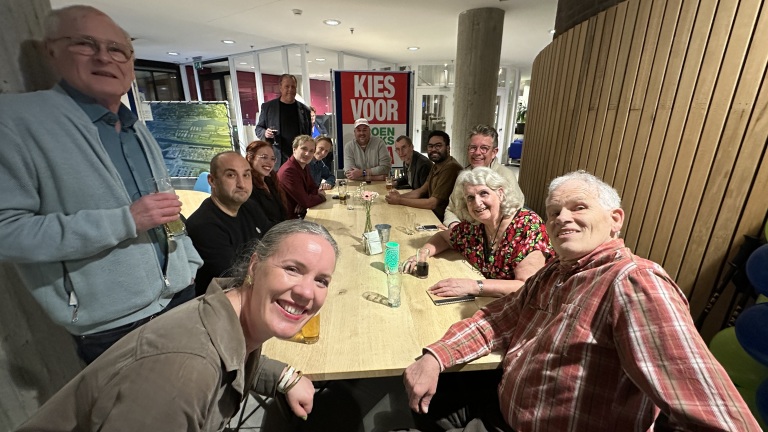 Mensen zitten rond een lange tafel in een kamer, lachend naar de camera, met een bord met de tekst "KIES VOOR" op de achtergrond.