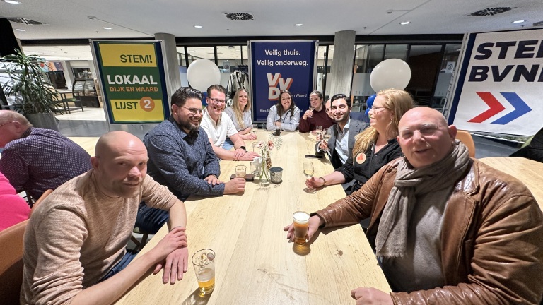 Mensen zitten rond een tafel met drankjes, omgeven door verkiezingsposters van verschillende politieke partijen.