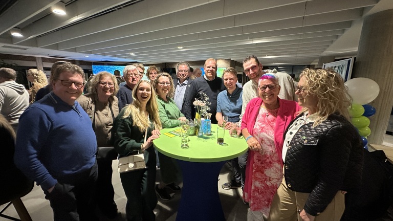 Groep mensen verzameld rond een tafel op een feestelijke bijeenkomst met kleurrijke ballonnen op de achtergrond.