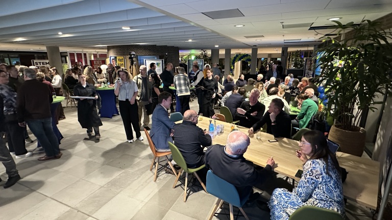 Een groep mensen op een bijeenkomst in een ruime, verlichte zaal met tafels en kleurrijke stoelen.