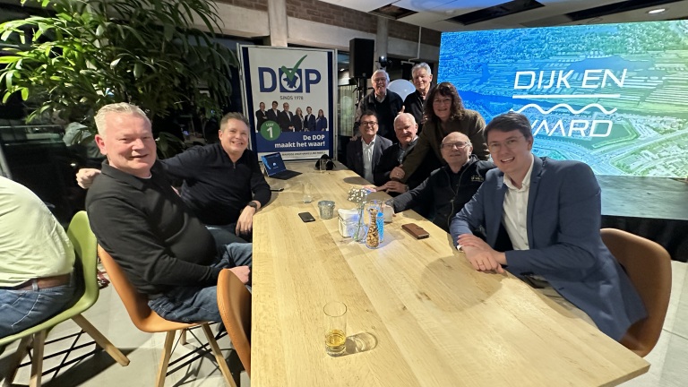 Groep mensen zit rondom een tafel in een kamer met een groot scherm en een poster van een politieke partij genaamd "DOP".