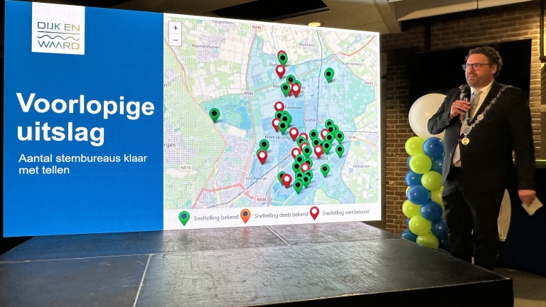 Een presentator staat naast een scherm met de tekst "Voorlopige uitslag" en een kaart met gemarkeerde locaties.