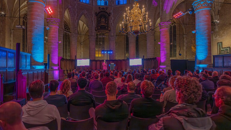 Een presentator spreekt voor een groot publiek in een historische kerk met kleurrijke verlichting en een kroonluchter.