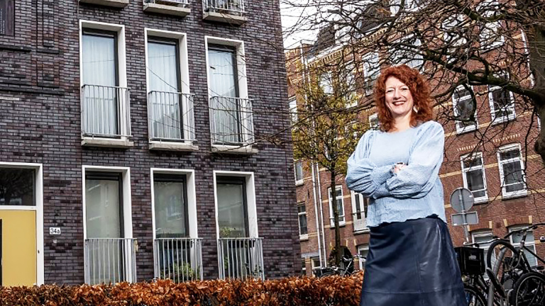 Vrouw met rood haar en blauwe trui staat glimlachend voor een bakstenen gebouw met gele deur en balkons.