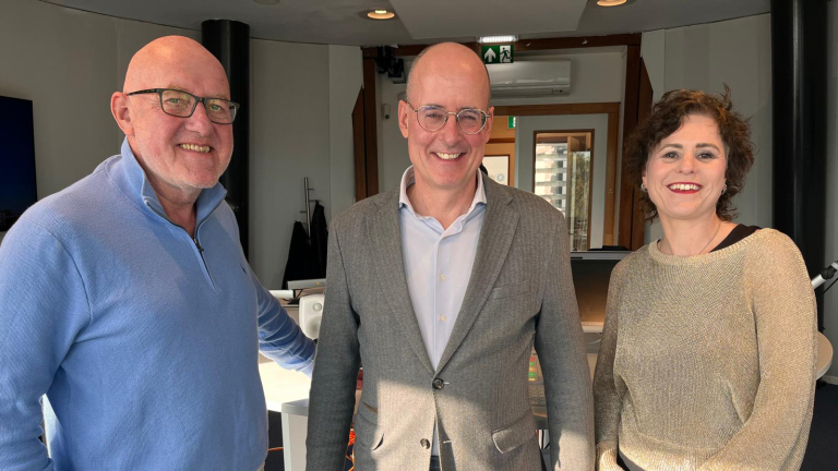 Drie glimlachende collega’s in kantoor, twee mannen en een vrouw, poseren voor de camera tussen bureaus en apparatuur