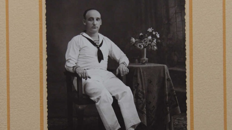 Zwart-wit foto van een man in een matrozenuniform die op een stoel zit naast een tafeltje met bloemen in een vaas.